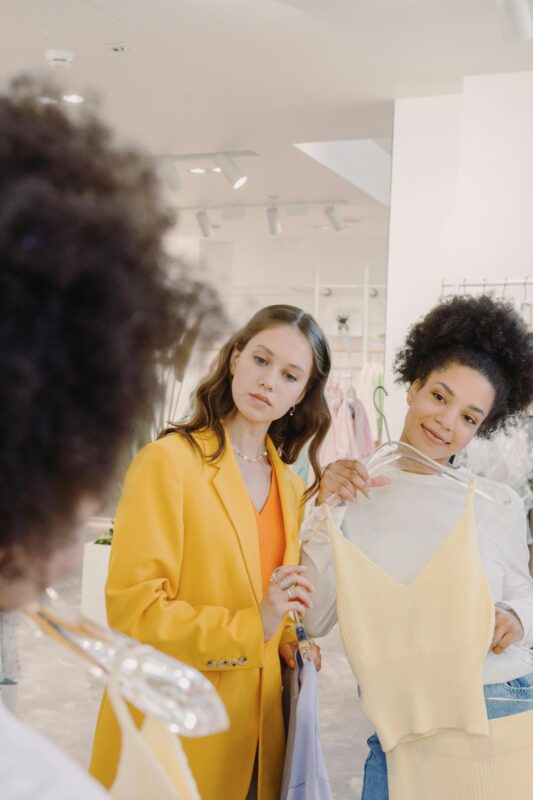 Two women shopping in a trendy boutique, trying on fashionable clothes with a mirror reflection.